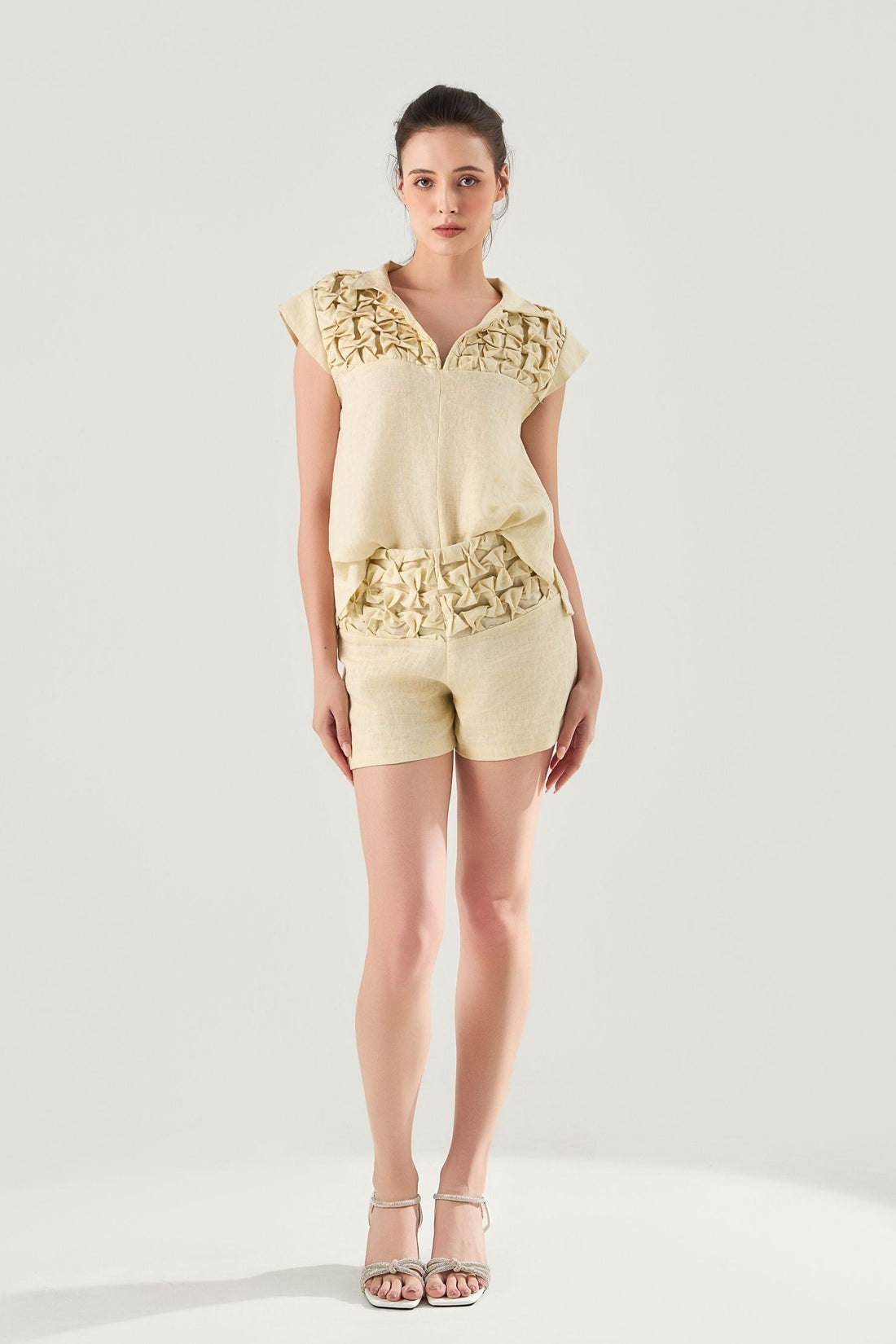 Woman wearing a light-colored Aria Hand-Smocked pure washed Linen top and shorts with interesting hand-smocking details on a white background